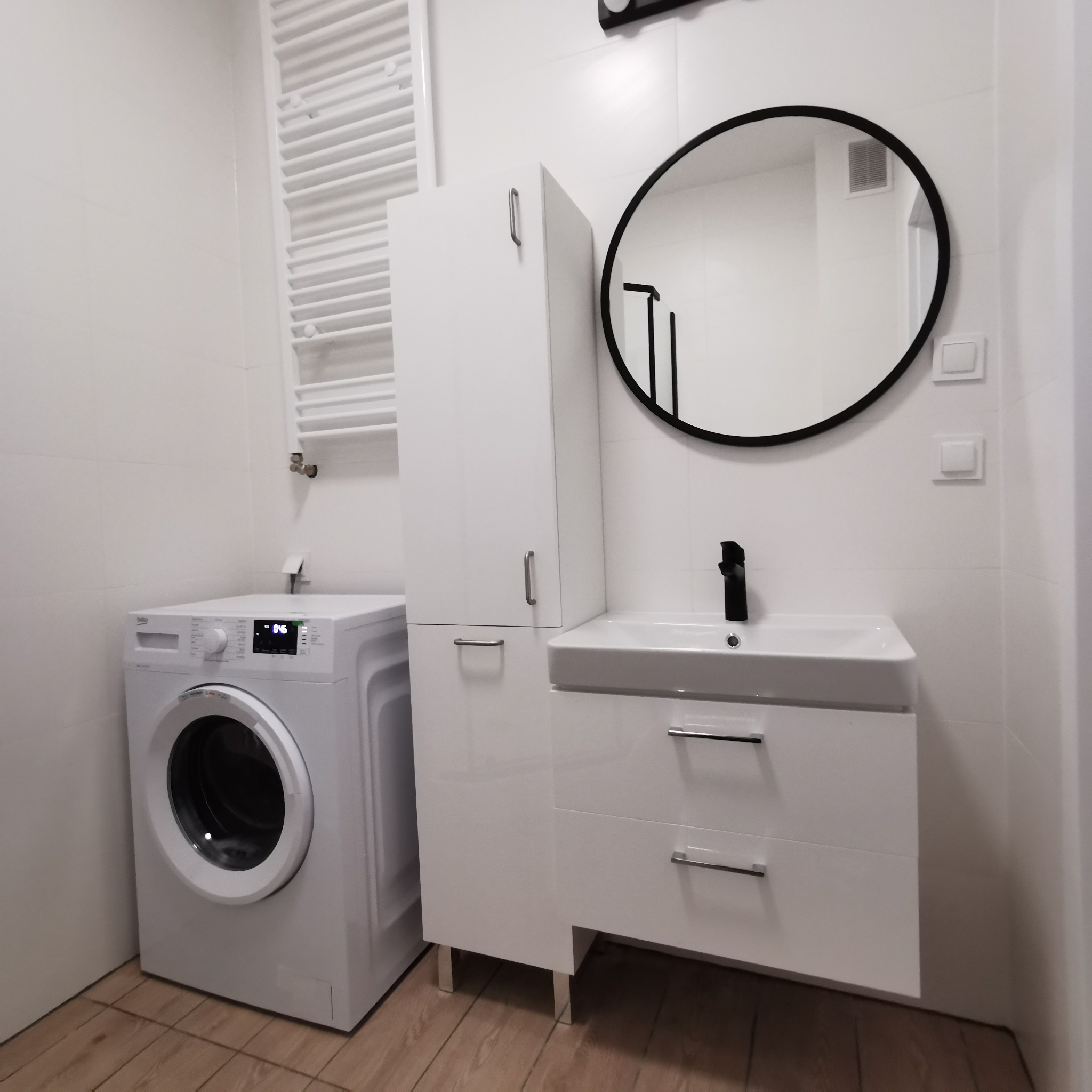 Biała łazienka z pralką Beko, szafką umywalkową z czarną armaturą, okrągłym lustrem w czarnej ramie i grzejnikiem drabinkowym na ścianie z białych płytek.