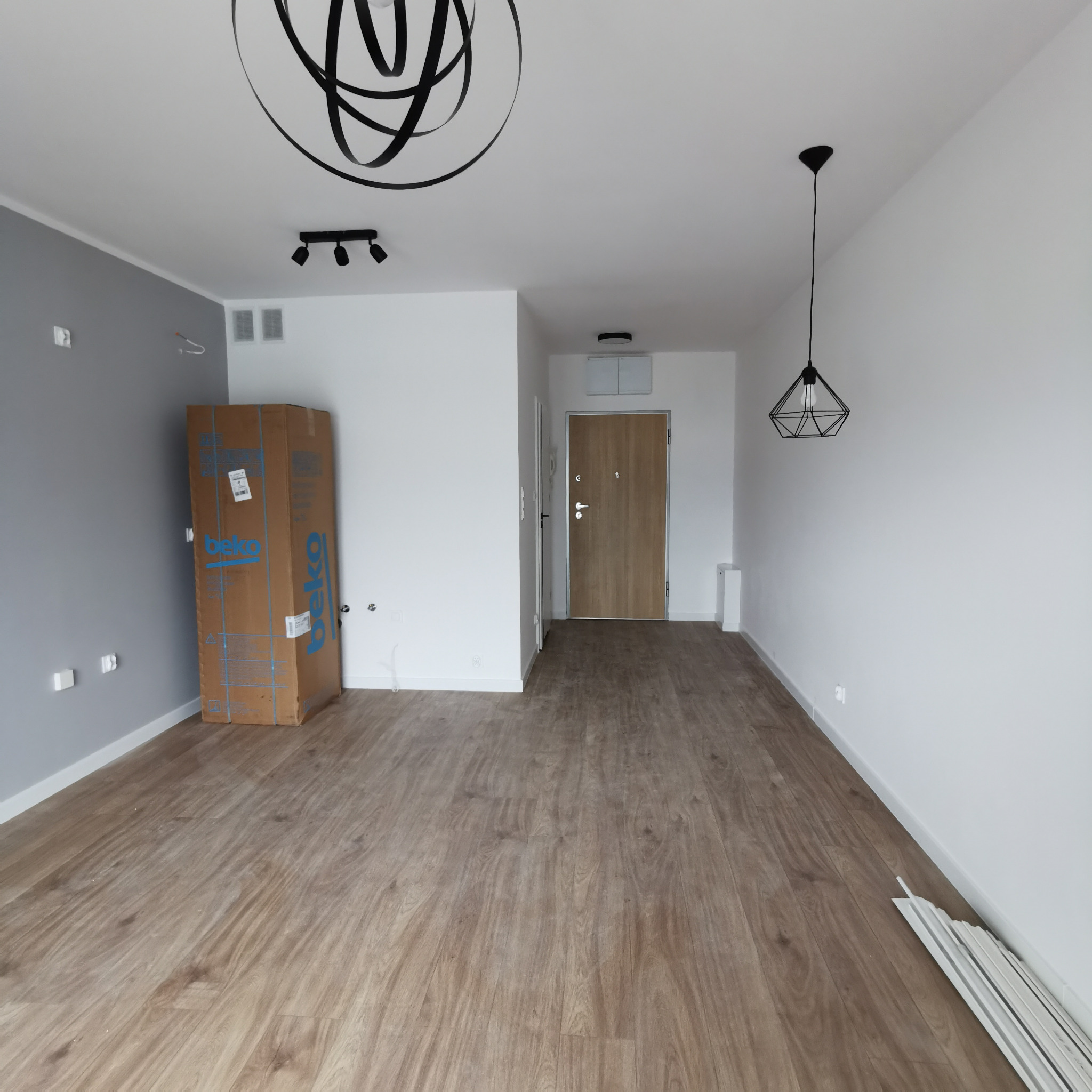 Pusty pokój po remoncie z drewnianą podłogą, szarą ścianą, modernistycznymi lampami i kartonem po sprzęcie AGD, oczekujący na umeblowanie.