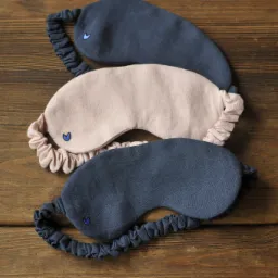 Trzy lniane maski do spania: dwie granatowe i jedna różowa, każda z małym, haftowanym niebieskim serduszkiem, leżą na drewnianym stole.