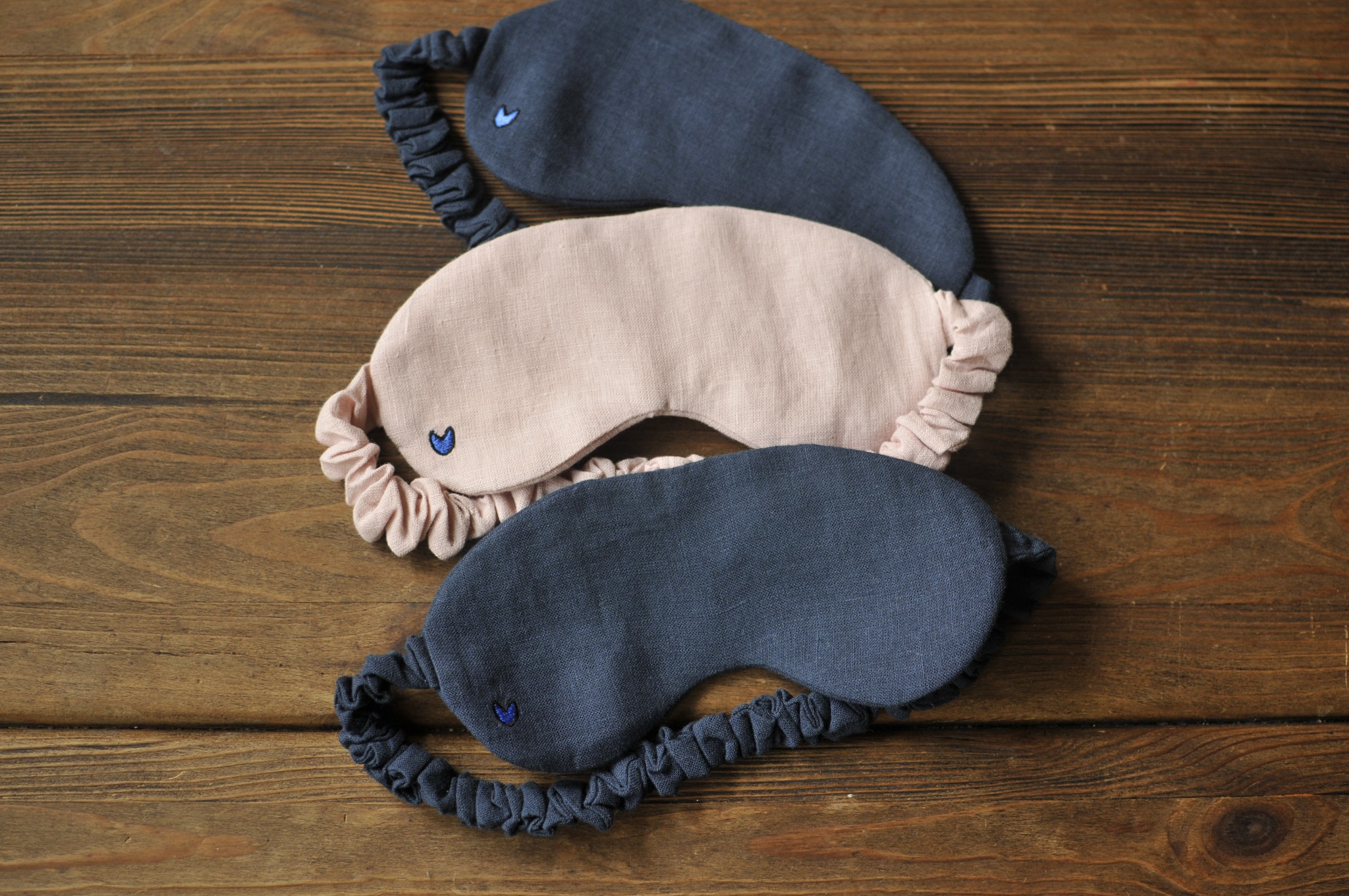 Trzy lniane maski do spania: dwie granatowe i jedna różowa, każda z małym, haftowanym niebieskim serduszkiem, leżą na drewnianym stole.