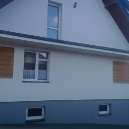 Dwukondygnacyjny dom z białą elewacją i fragmentami imitującymi drewno, szara podmurówka, okna z białymi ramami i firankami, czarne rynny, widok z zewnątrz.