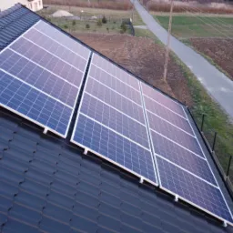 Panele słoneczne zamontowane na ciemnym, spadzistym dachu domu jednorodzinnego, widok z góry, w tle droga i pole.