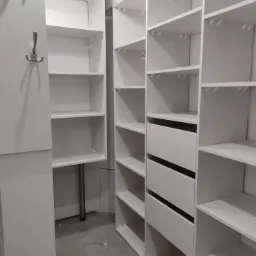 Biała, wnękowa garderoba z półkami i szufladami, oświetlona lampą sufitową z metalową siatką, z widocznymi wieszakami na ścianie po lewej stronie.