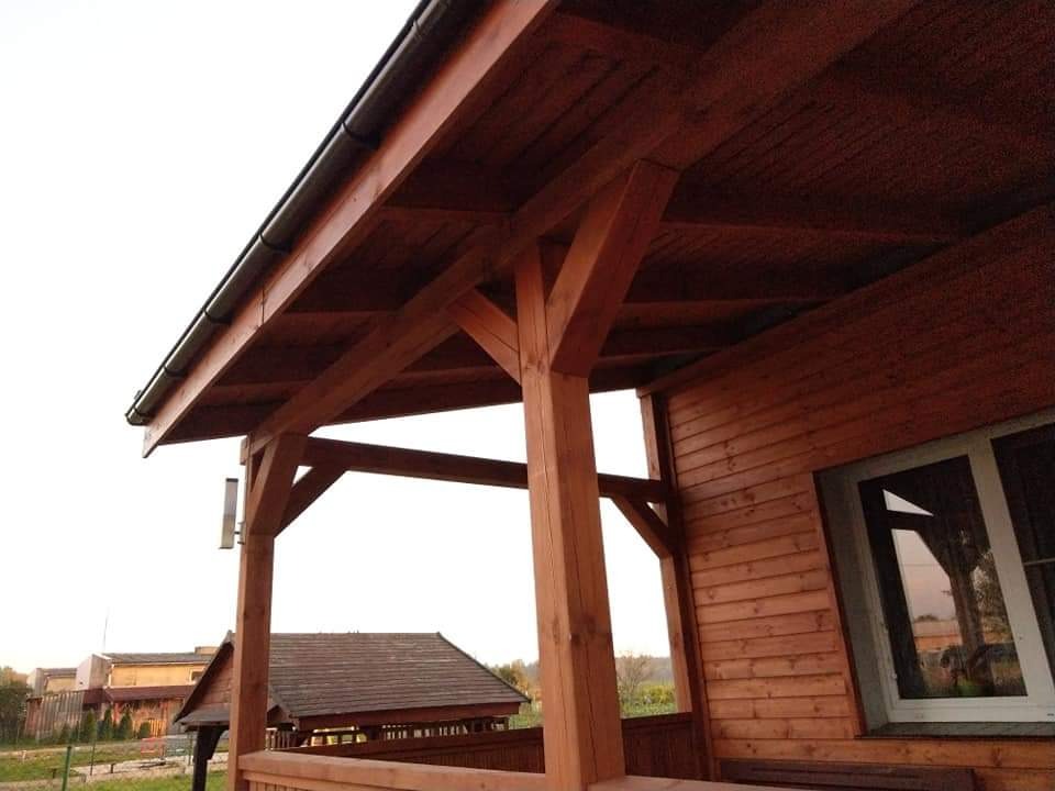 Drewniana altana z widoczną konstrukcją dachu, brązowymi belkami i deskami, z oknem w tle i fragmentem krajobrazu.