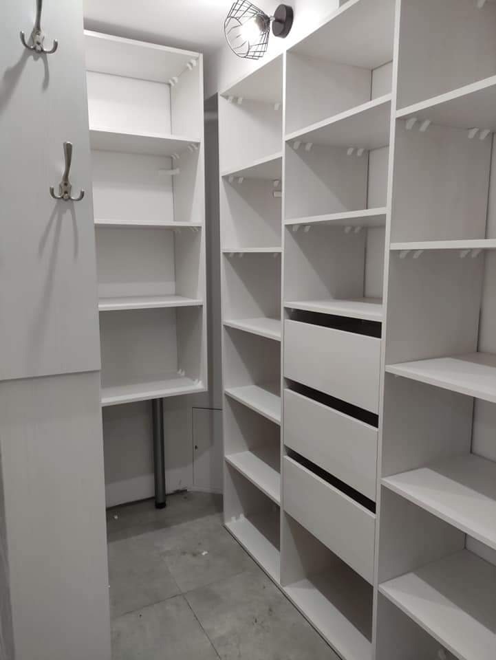 Biała, wnękowa garderoba z półkami i szufladami, oświetlona lampą sufitową z metalową siatką, z widocznymi wieszakami na ścianie po lewej stronie.