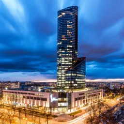 Wieczorny widok na Sky Tower w Łodzi, z długą ekspozycją świateł ulicznych i dynamicznymi chmurami na niebie.