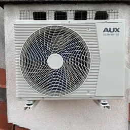 Zewnętrzna jednostka klimatyzacji AUX C-Smart zamontowana na elewacji budynku z widoczną kratką wentylacyjną powyżej.