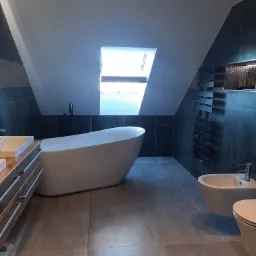 Nowoczesna łazienka na poddaszu z wolnostojącą wanną, czarną armaturą i grzejnikiem drabinkowym na tle grafitowych płytek, doświetlona oknem dachowym.