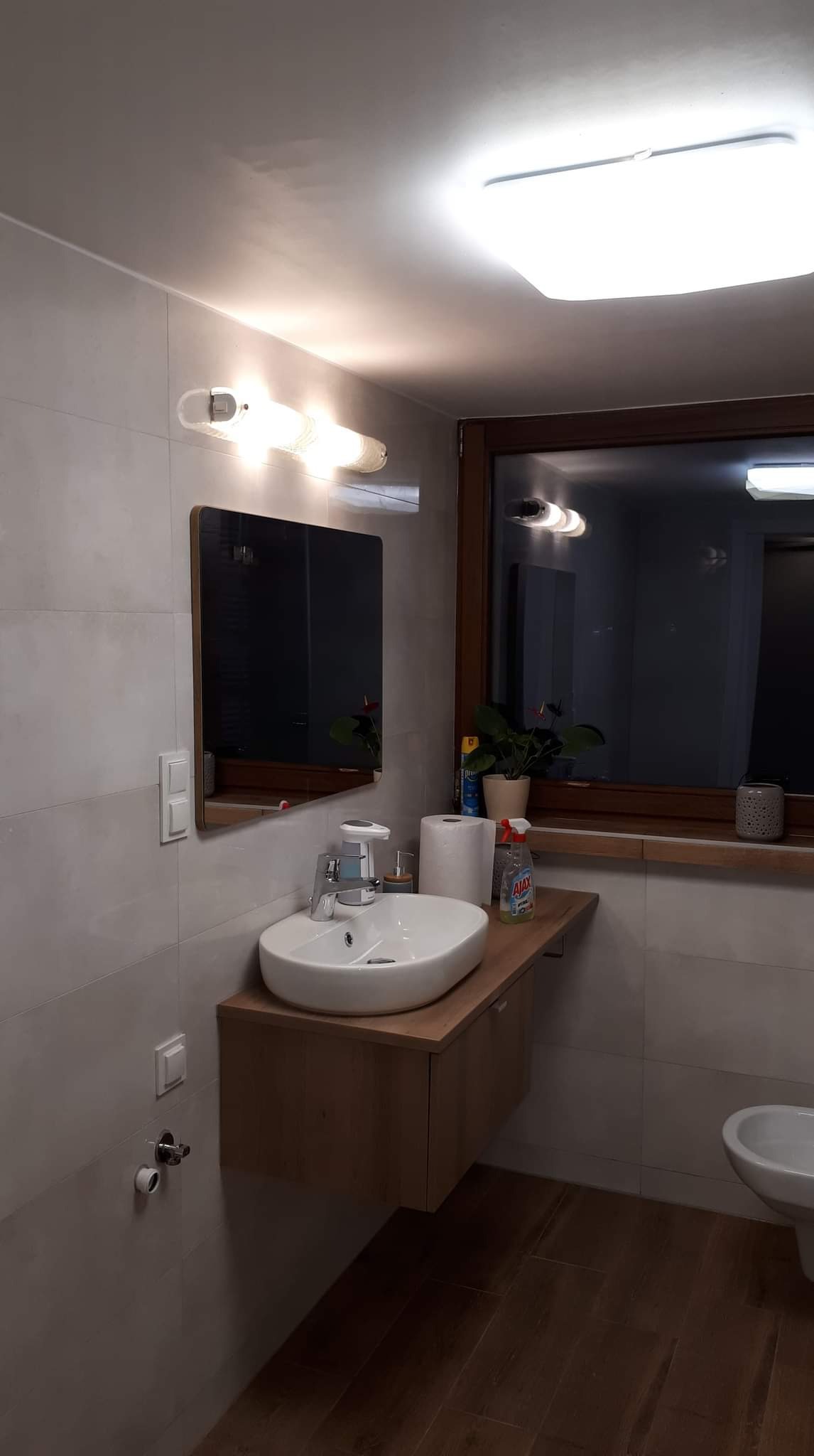 Nowoczesna łazienka z owalną umywalką nablatową na drewnianej szafce, szare płytki na ścianach, drewniana rama okna z rośliną w doniczce i widoczną toaletą w tle.