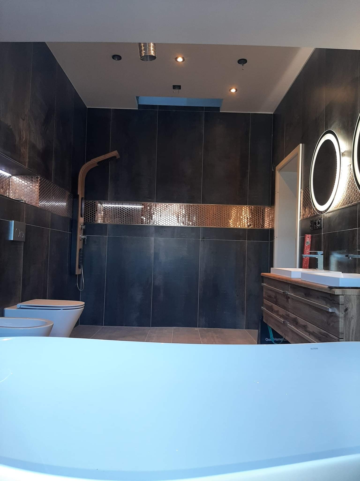 Nowoczesna łazienka z ciemnymi płytkami, mozaiką w kolorze miedzi, podświetlanymi lustrami i drewnianą szafką pod umywalką, widok zza wanny.