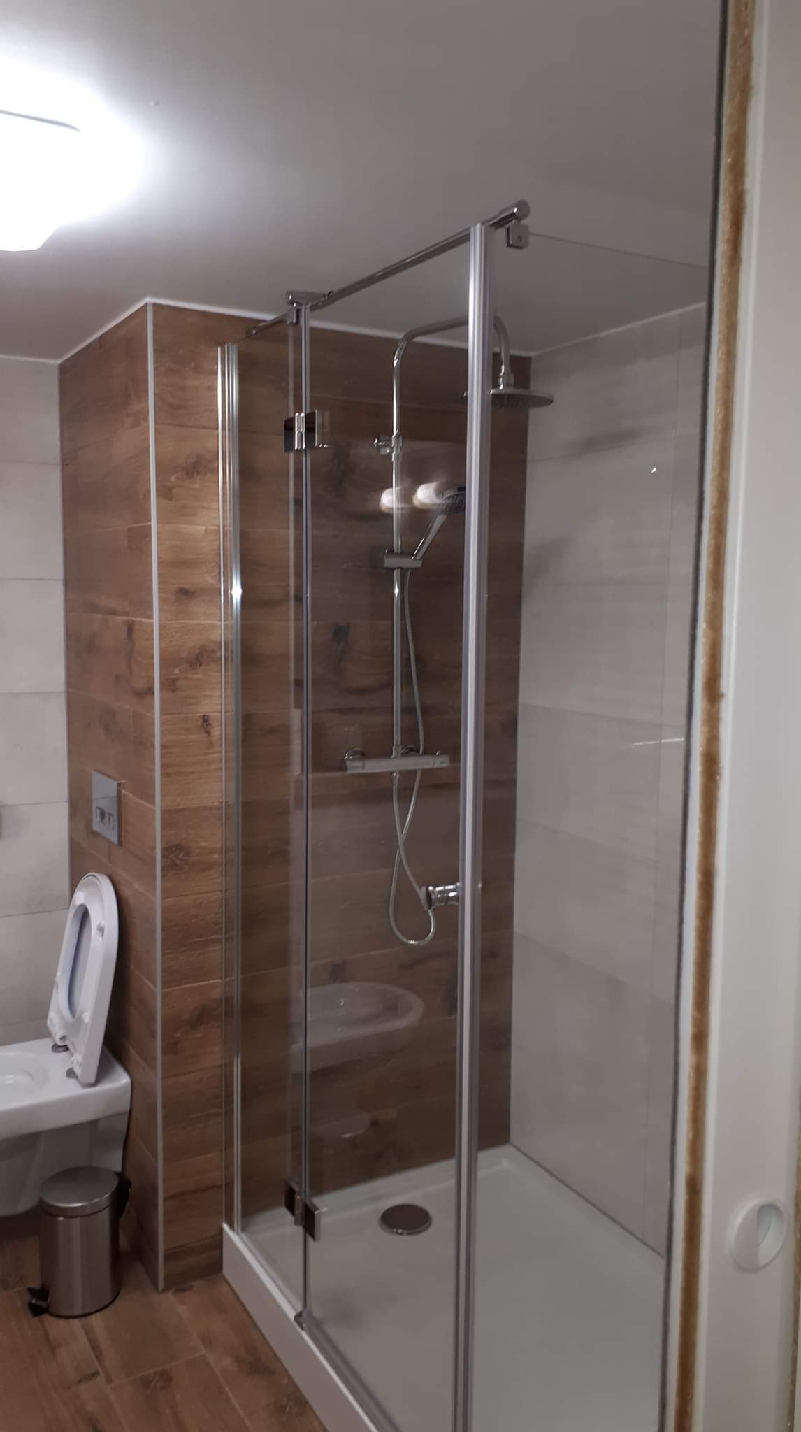 Kabina prysznicowa z chromowanymi detalami, widoczna umywalka i toaleta w łazience z płytkami imitującymi drewno.