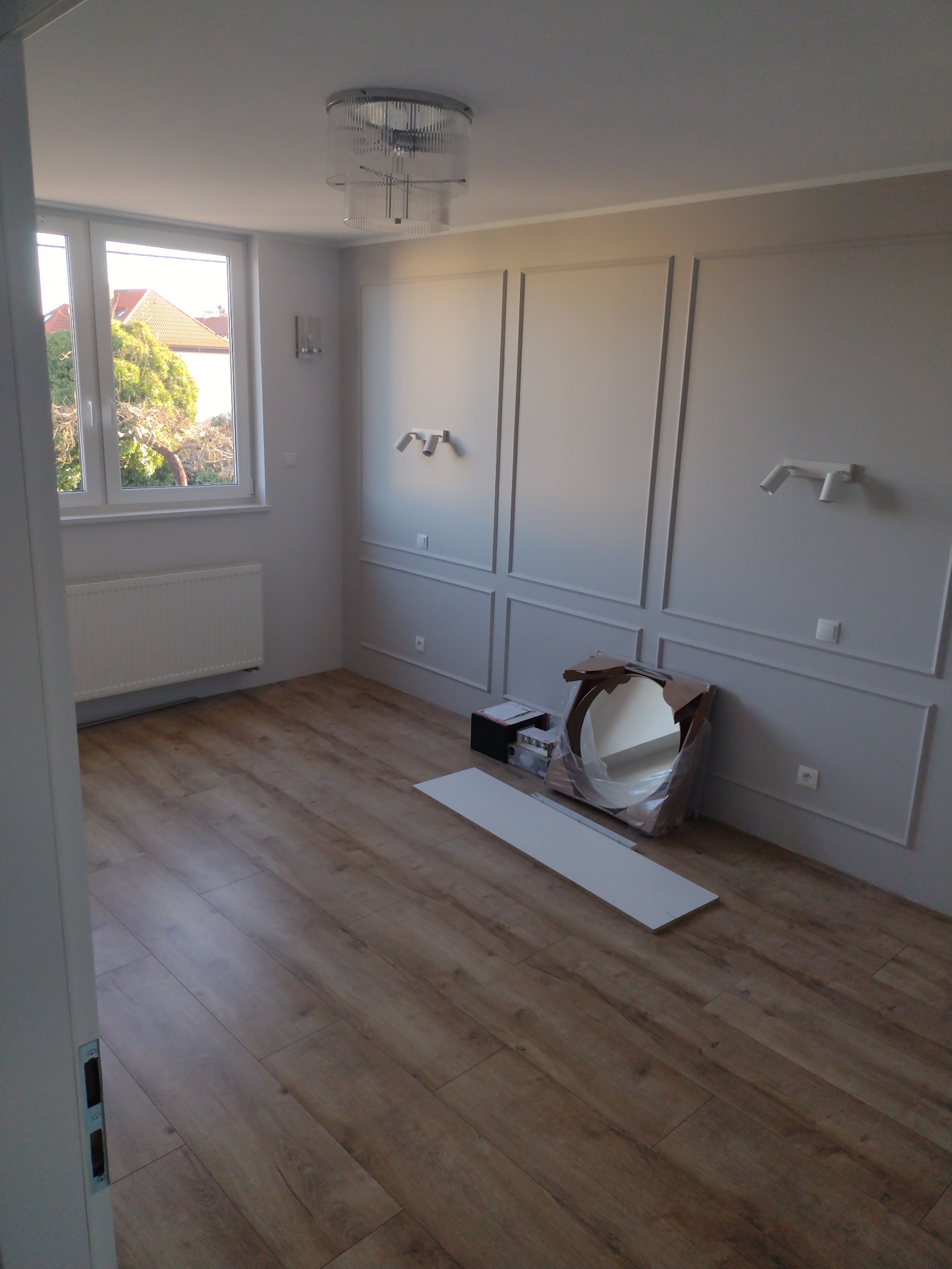 Pusty pokój z panelami podłogowymi, grzejnikiem, dekoracyjnymi listwami na ścianach, nowoczesnym oświetleniem sufitowym i ściennym, z lustrem w kartonie i listwą przypodłogową przygotowanymi...