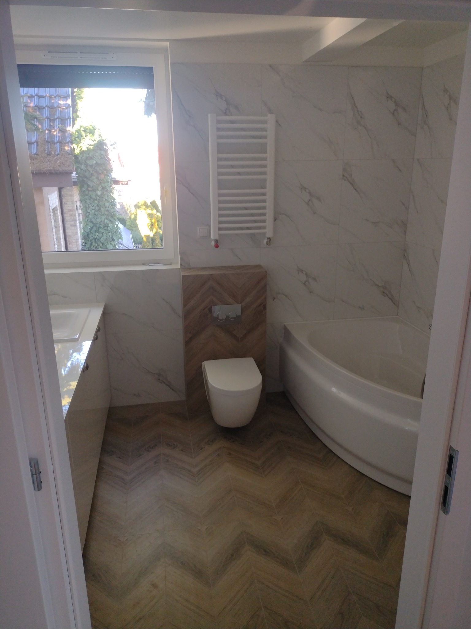 Wykończona łazienka z podłogą ułożoną w jodełkę, białą armaturą i marmurowymi płytkami na ścianach, widok zza uchylonych drzwi.