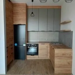 Nowoczesna kuchnia z frontami w kolorze szarym i drewnopodobnym, czarna lodówka w zabudowie, drewniana podłoga, minimalistyczne oświetlenie.