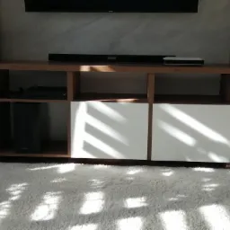 Zabudowa RTV z drewnianymi półkami i białymi frontami szafek, na tle ściany z efektem betonu, z widocznym sprzętem audio-video i puszystym, białym dywanem.