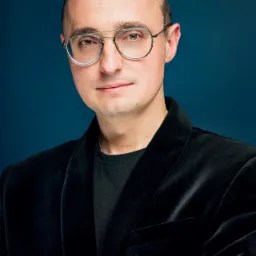 Portret mężczyzny w okularach i czarnym welurowym żakiecie na ciemnoniebieskim tle, z rękami skrzyżowanymi na piersiach.