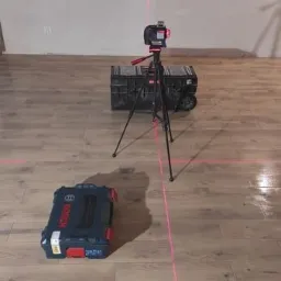 Poziomica laserowa ustawiona na podłodze z paneli, wyznaczająca linie, obok skrzynki narzędziowe Bosch.