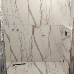 Glazur-Mac - Ściany i podłoga wyłożone białymi płytkami imitującymi marmur z brązowymi żyłami, widoczne otwory na armaturę i fragment niezabezpieczonej ściany.