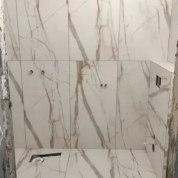 Ściany i podłoga wyłożone białymi płytkami imitującymi marmur z brązowymi żyłami, widoczne otwory na armaturę i fragment niezabezpieczonej ściany.