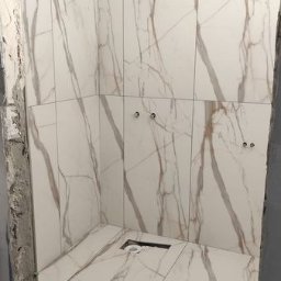 Glazur-Mac - Płytki imitujące marmur w kabinie prysznicowej w trakcie montażu, widoczne otwory na armaturę i niedokończone wykończenie ściany.