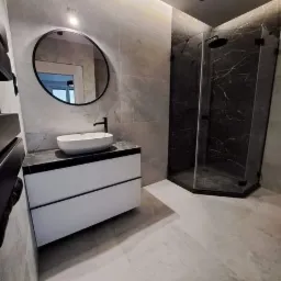 Nowoczesna łazienka w odcieniach szarości z kabiną prysznicową z czarnymi elementami, okrągłym lustrem z czarną ramą nad białą szafką z umywalką i podświetleniem LED na suficie.