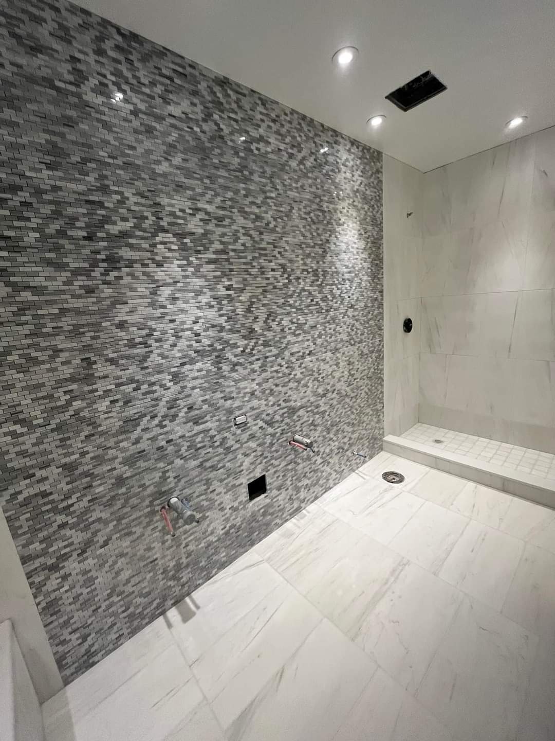 Łazienka w trakcie wykańczania: ściana pokryta mozaiką w odcieniach szarości, białe płytki imitujące marmur na podłodze i części ściany prysznicowej, widoczne wyprowadzenia instalacji wodnej...