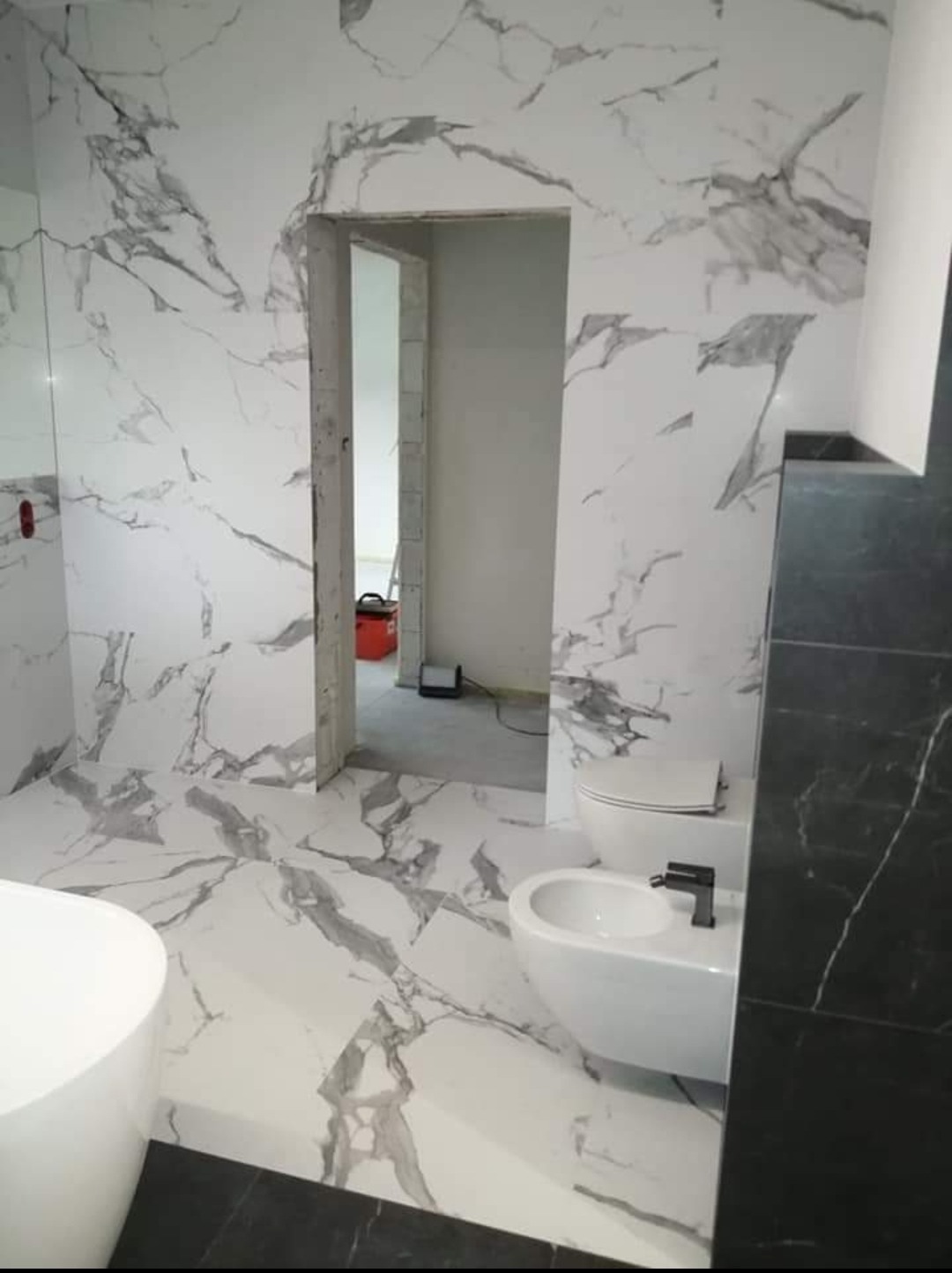 Łazienka w trakcie wykańczania z białymi płytkami imitującymi marmur na ścianach i podłodze, widoczna toaleta, bidet z czarną armaturą oraz fragment wanny, w tle otwarte przejście do innego...