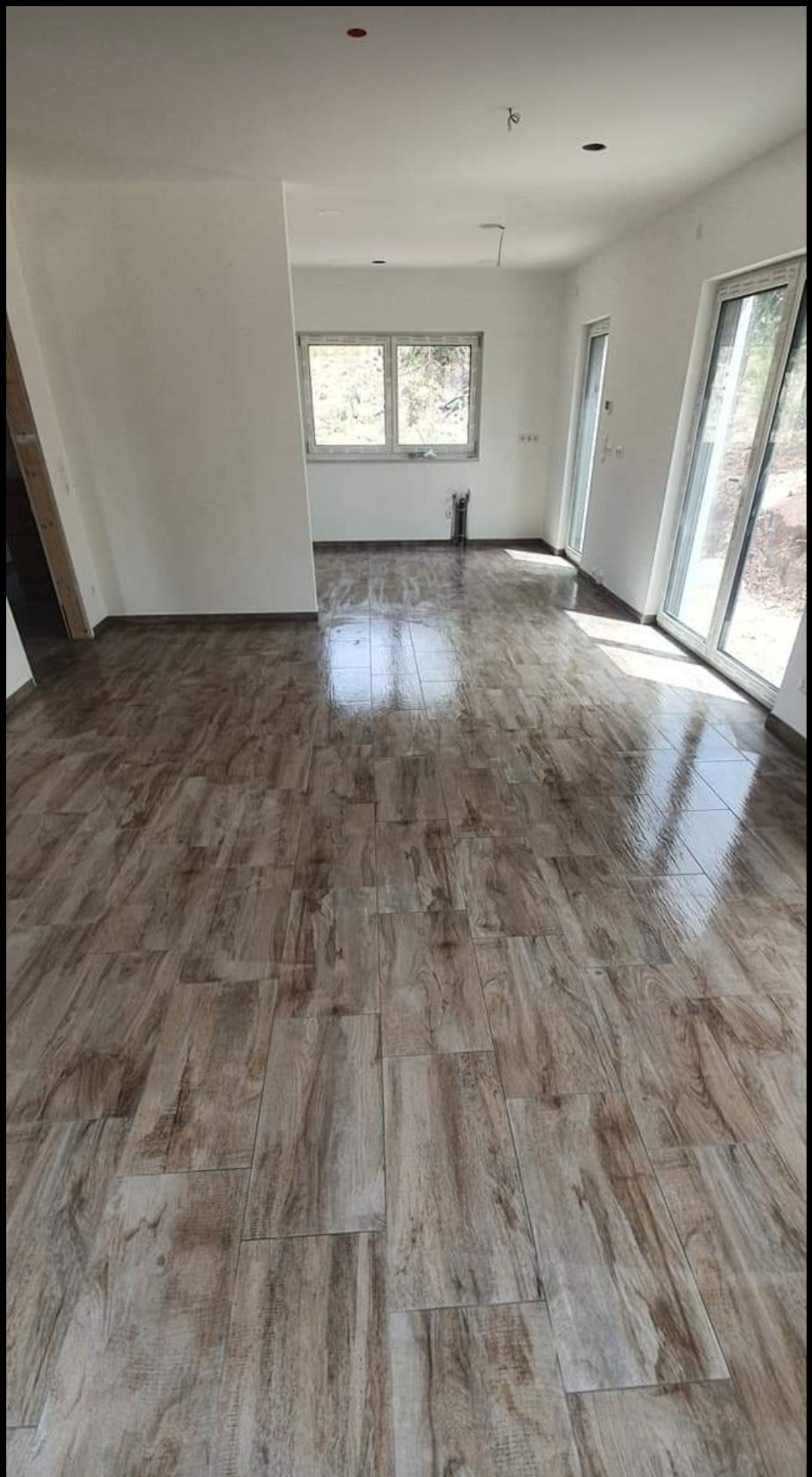 Ułożona podłoga z płytek imitujących drewno w jasnym, przestronnym pokoju z dużymi oknami balkonowymi, widoczne niedoskonałości na ścianach sugerujące świeżo zakończony remont.