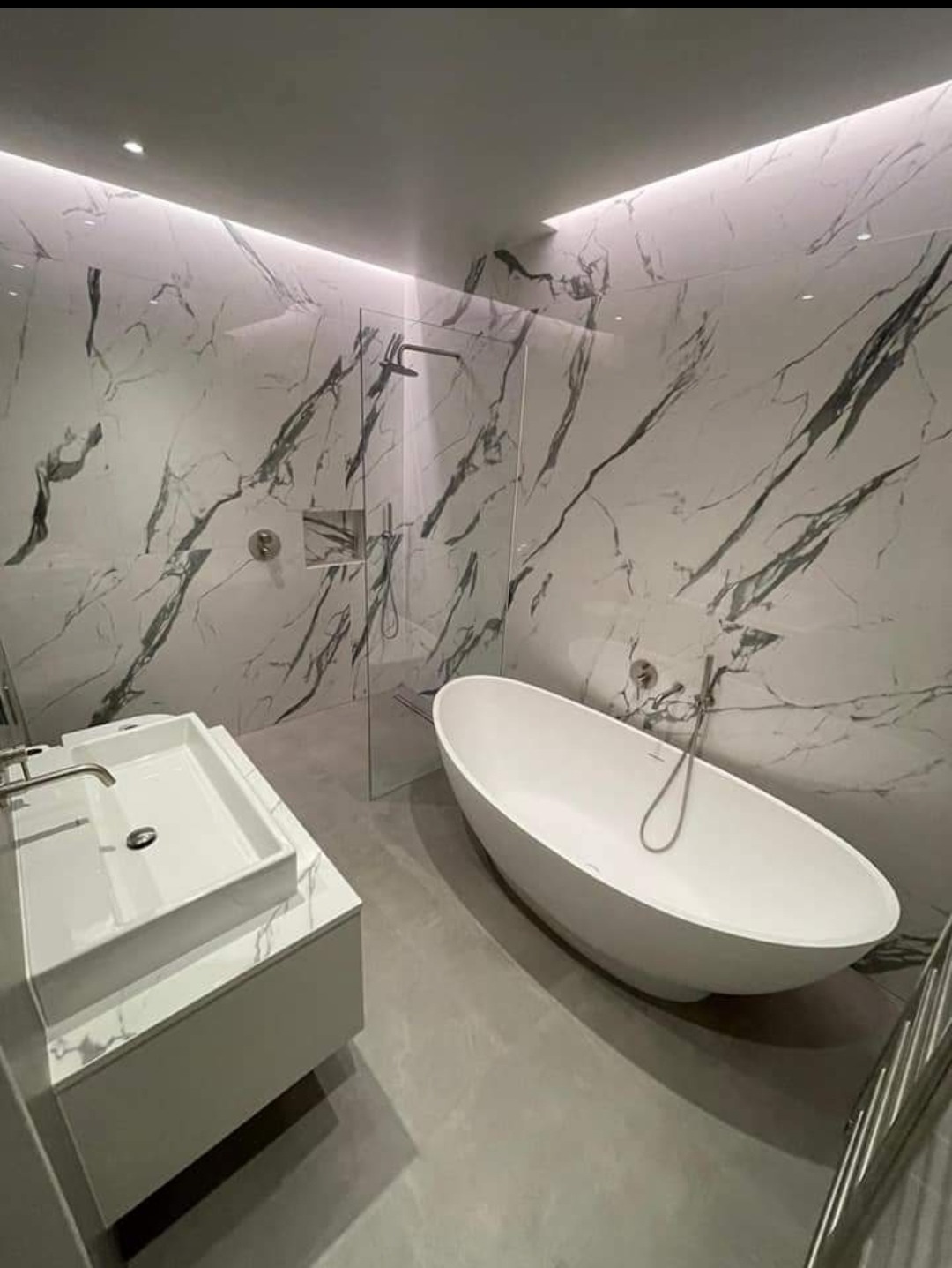 Nowoczesna łazienka z marmurowymi płytkami na ścianach, wolnostojącą białą wanną, szklaną kabiną prysznicową i umywalką nablatową.