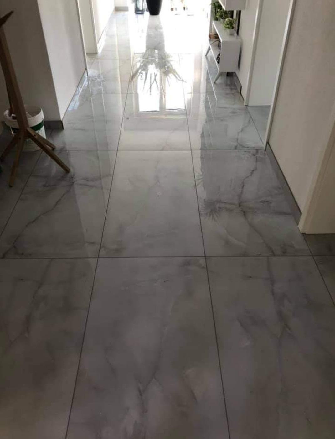 Błyszcząca, szara marmurowa podłoga z płytek w korytarzu, z widocznym odbiciem okna i roślin doniczkowych, oraz wiadro z farbą i drewniany stojak malarski w rogu.