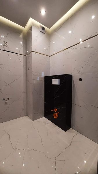 Wykończona łazienka z białymi płytkami imitującymi marmur, czarną zabudową stelaża WC z białym przyciskiem, podświetleniem LED na suficie i złotymi listwami dekoracyjnymi.