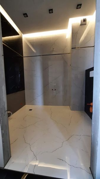 Nowoczesna łazienka w trakcie wykańczania, z białymi płytkami imitującymi marmur na podłodze i ścianach, oświetlenie LED w suficie, widoczne fragmenty instalacji hydraulicznej.