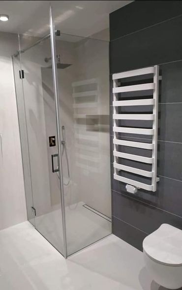 Nowoczesna kabina prysznicowa ze szklanymi drzwiami, chromowaną armaturą i odpływem liniowym, obok biały grzejnik drabinkowy na szarej ścianie z płytek, widoczny fragment toalety.