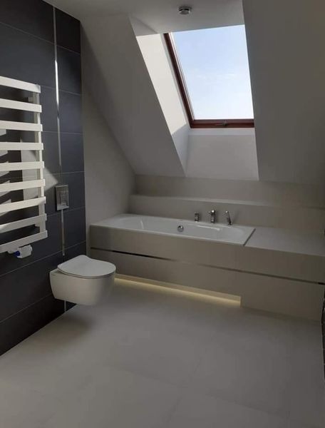 Nowoczesna łazienka na poddaszu z oknem dachowym, białą armaturą i szarą ścianą z grzejnikiem drabinkowym.