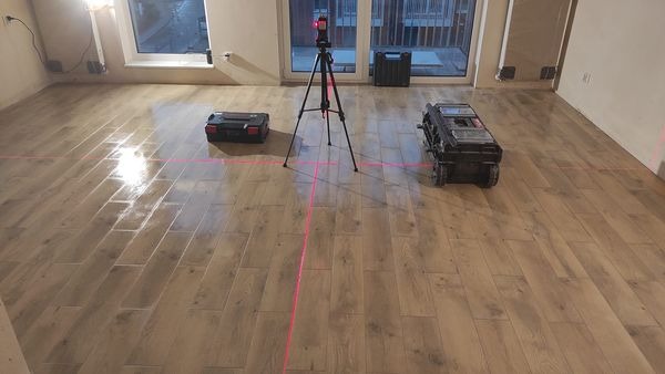Układanie podłogi z płytek imitujących drewno, z użyciem lasera krzyżowego do precyzyjnego wyznaczania linii, widoczne narzędzia i akcesoria glazurnicze.
