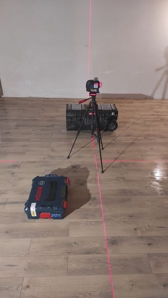 Poziomica laserowa ustawiona na podłodze z paneli, wyznaczająca linie, obok skrzynki narzędziowe Bosch.