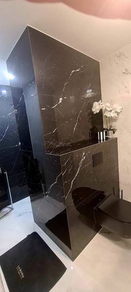 Nowoczesna łazienka z czarną marmurową obudową toalety, białą marmurową podłogą i ścianami, czarną miską ustępową i czarnym dywanikiem z logo 'La Mania'.