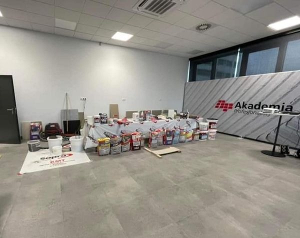 Sala konferencyjna z przygotowanymi materiałami budowlanymi, w tym farbami, gładziami i zaprawami, rozłożonymi na stole i podłodze, z widocznym banerem 'Akademia Profesjonalna' w tle. Przygotowania...