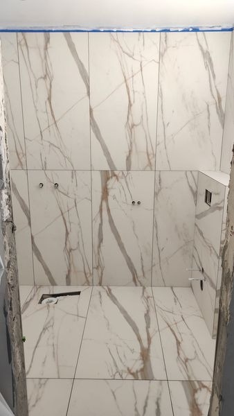 Ściany i podłoga wyłożone białymi płytkami imitującymi marmur z brązowymi żyłami, widoczne otwory na armaturę i fragment niezabezpieczonej ściany.