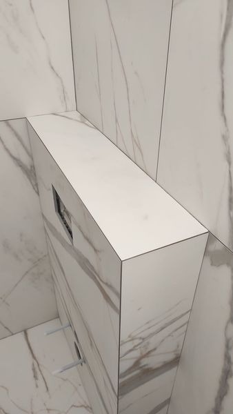 Obudowa stelaża podtynkowego WC wykończona płytkami imitującymi biały marmur z szarymi żyłami, dopasowanymi do płytek na ścianie i podłodze, widoczne elementy montażowe.