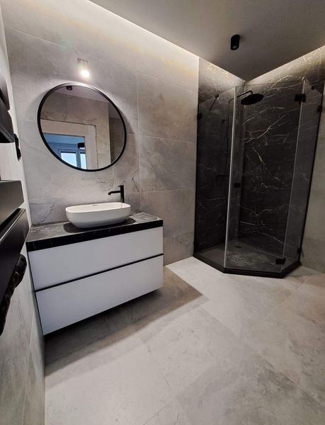 Nowoczesna łazienka w odcieniach szarości z kabiną prysznicową z czarnymi elementami, okrągłym lustrem z czarną ramą nad białą szafką z umywalką i podświetleniem LED na suficie.