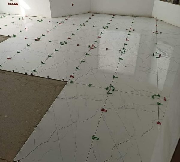 Układanie białych płytek podłogowych z efektem marmuru, z użyciem klipsów poziomujących w kolorach zielonym i czerwonym, widoczne niedokończone fragmenty podłogi.
