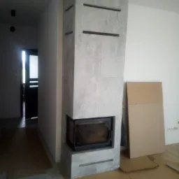 Murowany kominek z szarej okładziny, czarny wkład kominkowy, zabezpieczony kartonem na podłodze, widok na drzwi i fragment ściany.