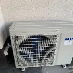 Zewnętrzna jednostka klimatyzacji AUX zamontowana na szarej kostce brukowej przy ścianie budynku z widocznym logo i naklejką informacyjną firmy instalacyjnej.