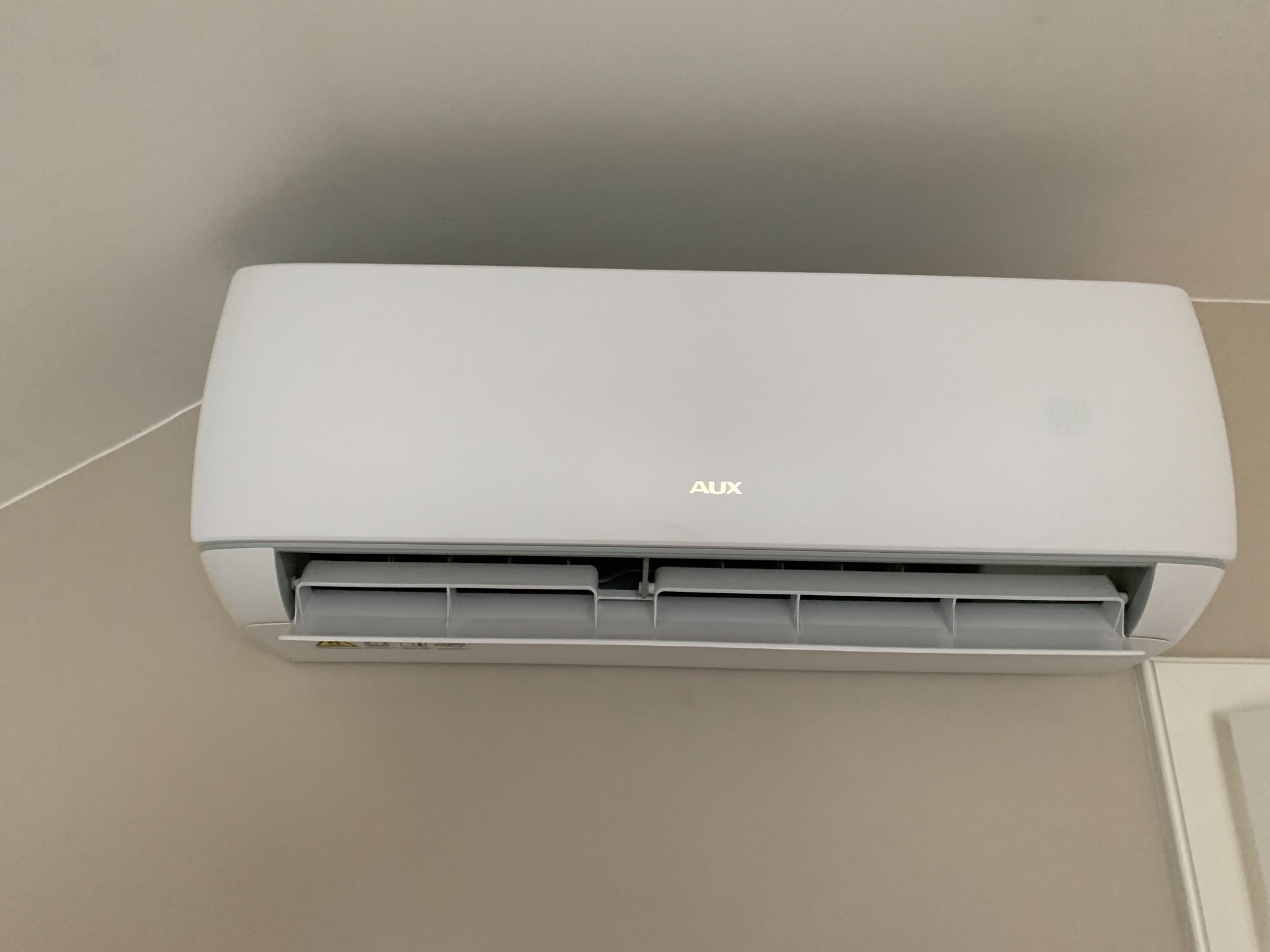 Biała klimatyzacja ścienna marki AUX, zamontowana w pomieszczeniu o jasnych ścianach, widoczne żaluzje wylotu powietrza.