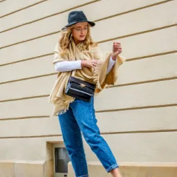 Kobieta w okularach, kapeluszu i beżowym ponczo z frędzlami, pozująca z czarną torebką na łańcuszku, dżinsach i białych sneakersach na tle kremowej ściany.