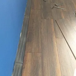 Fragment podłogi z ciemnobrązowych paneli ułożonych przy niebieskiej ścianie, widoczne narzędzia montażowe i listwa przypodłogowa.