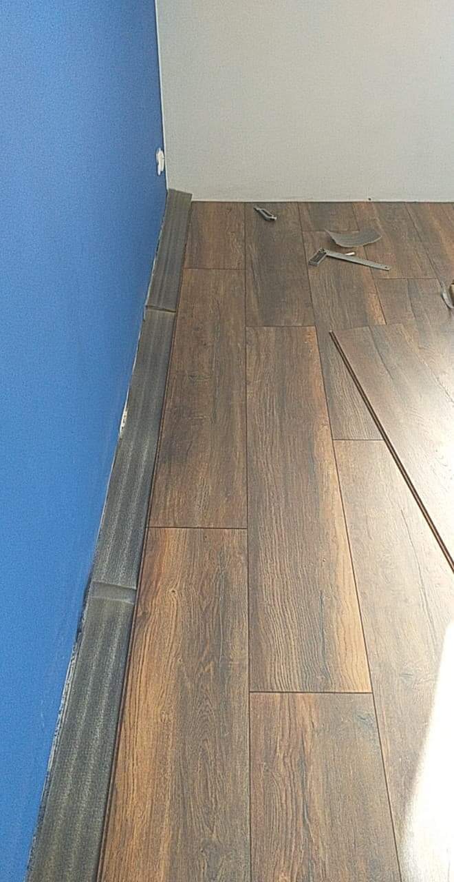 Fragment podłogi z ciemnobrązowych paneli ułożonych przy niebieskiej ścianie, widoczne narzędzia montażowe i listwa przypodłogowa.