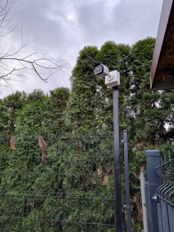 Zewnętrzna kamera monitoringu zamontowana na czarnym słupku przy ogrodzeniu z siatki, na tle zielonych drzew iglastych i pochmurnego nieba.