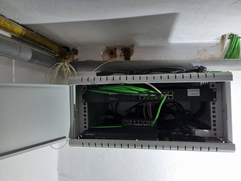 Otwarta szafa rack z zainstalowanymi urządzeniami sieciowymi, widoczne kable w kolorze zielonym i czarnym, oraz wystające przewody ze ściany i rury pod sufitem.